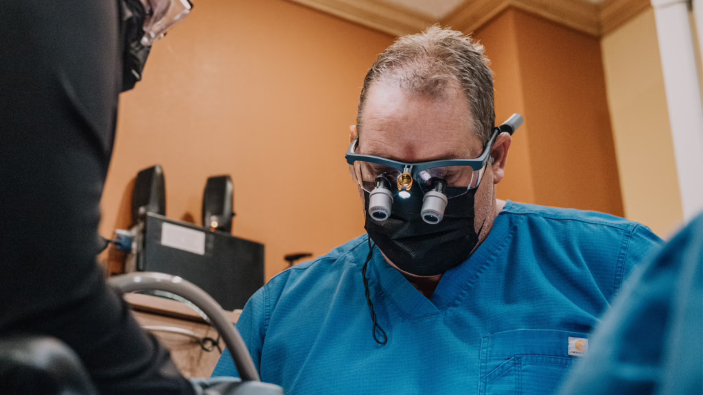 Surgeon wearing head-mounted magnifying loupes and a black mask, focusing on a patient during a procedure in blue scrubs.
