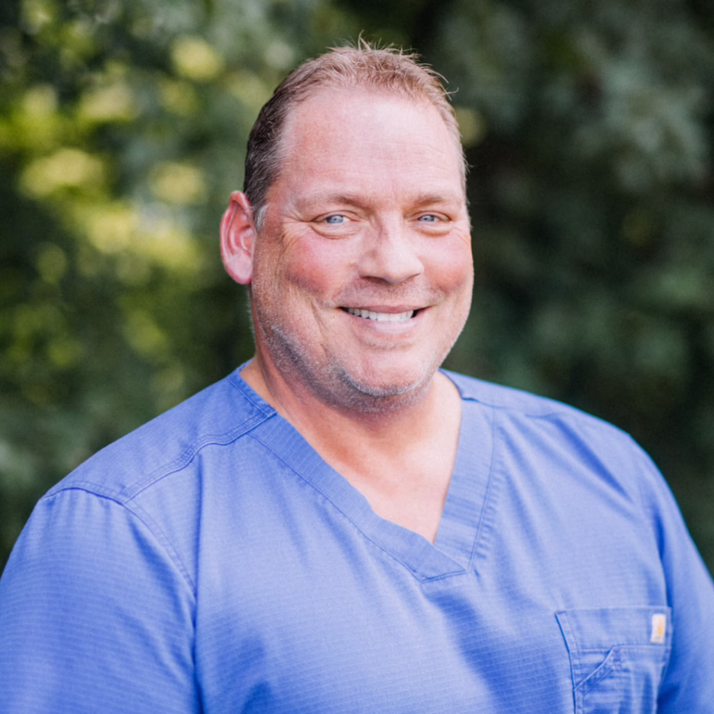 Smiling man in blue scrubs standing outdoors with green foliage in background.