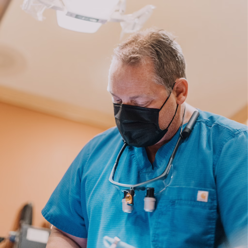 Surgeon in blue scrubs wearing a black face mask and loupes around the neck, focused on a procedure in an operating room.
