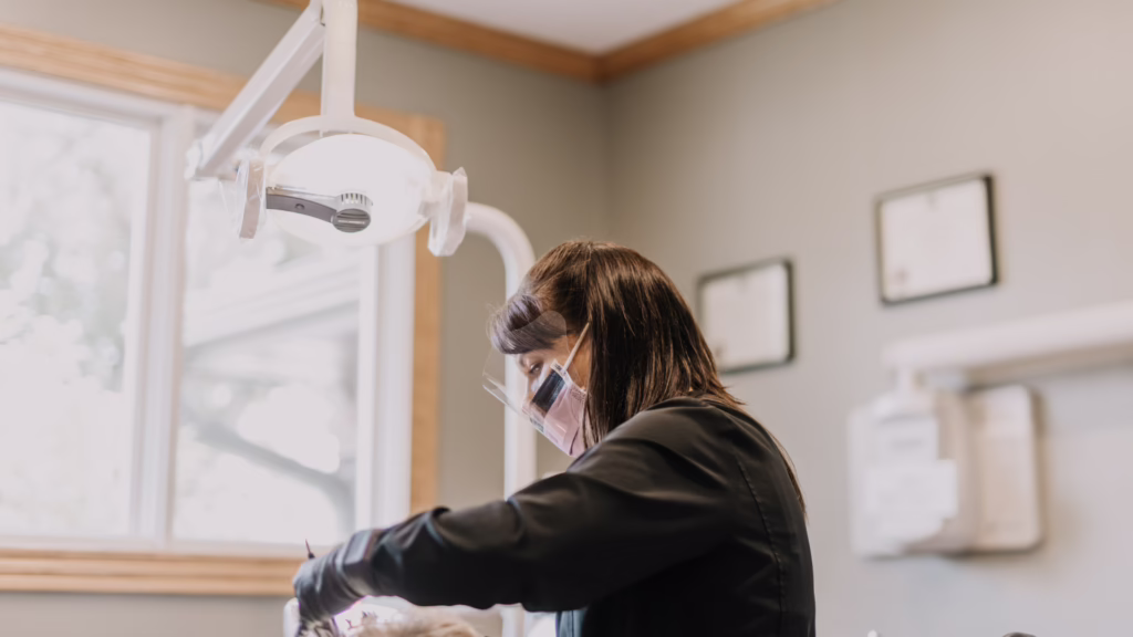 Dental hygienist wearing a mask and gloves performing a procedure under an overhead light in a clinic