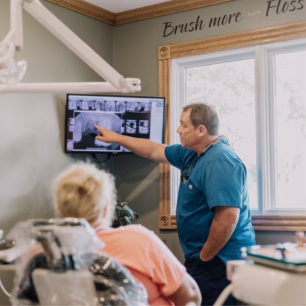 Dentist in blue scrubs points to a dental X-ray on a monitor while explaining to a patient in a dental chair.