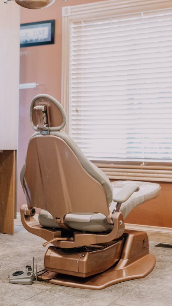 Brown reclining dental chair in a clinic treatment room with window blinds behind it and a foot pedal visible at floor level.