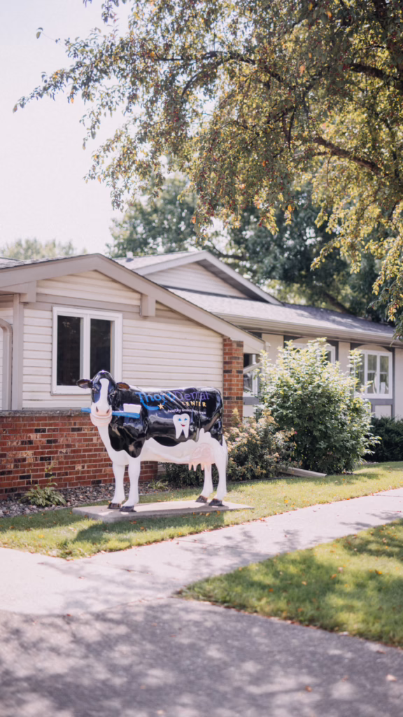 Life-size cow statue in a front yard with a sign for a dental center, in a suburban neighborhood.