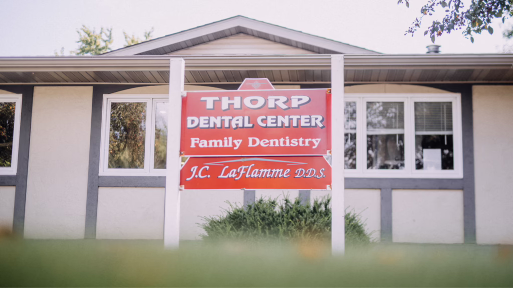Red sign for Thorp Dental Center advertising Family Dentistry in front of a small office building, with windows and beige siding in the background, and a green hedge below.